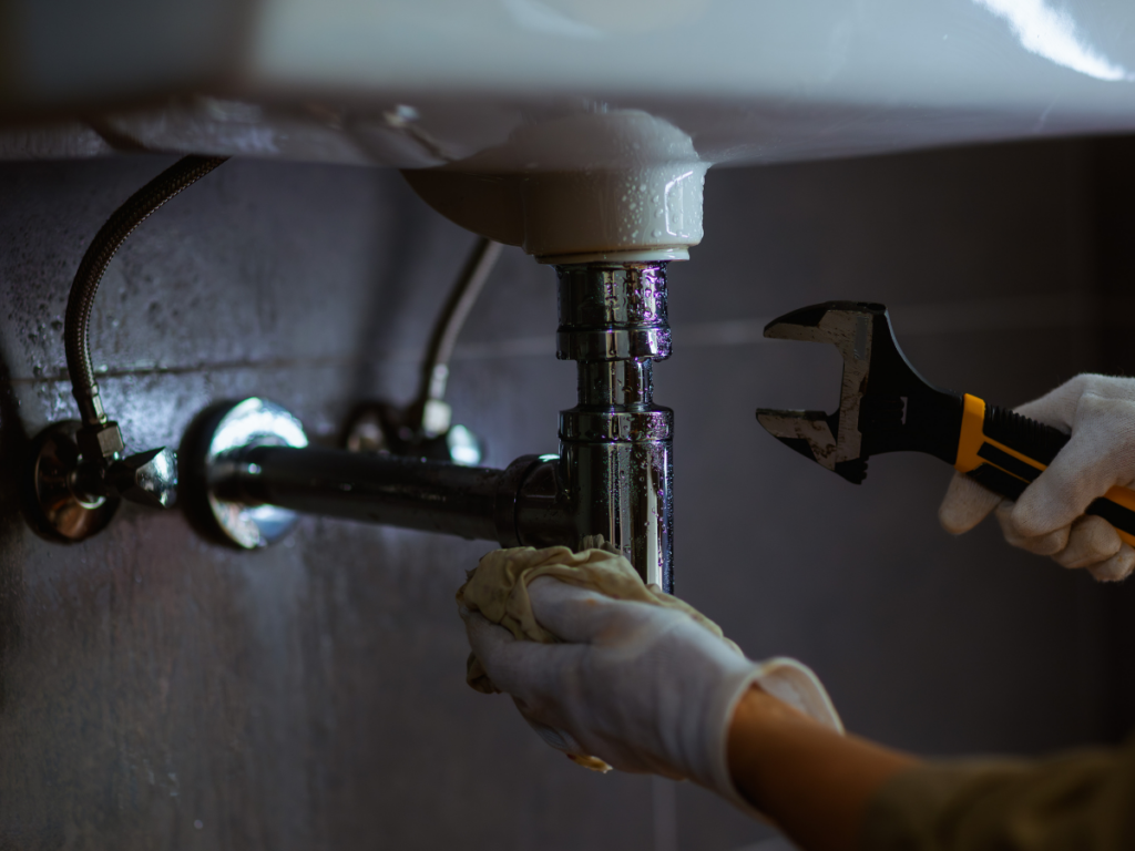 Professional plumber using a wrench to tighten a leaking P-trap pipe under a bathroom pedestal sink.
