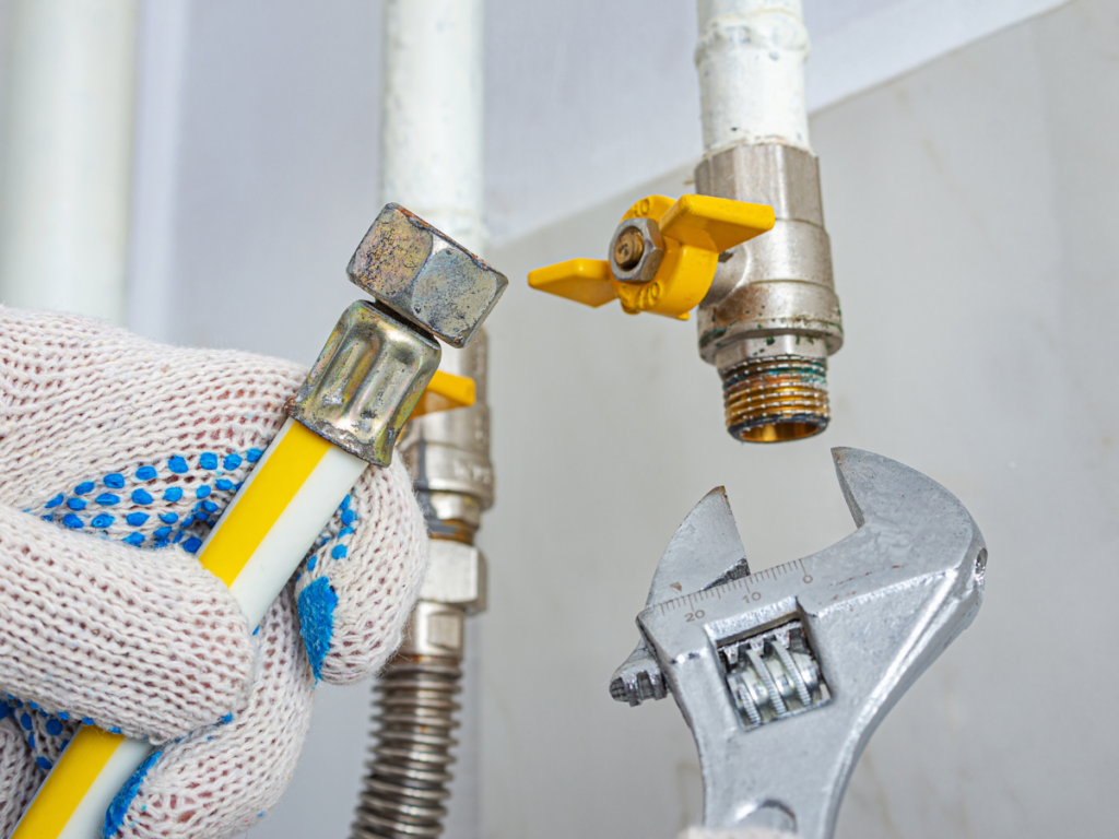 A-Comfort Services technician in safety gloves preparing to connect a yellow-jacketed flexible gas line to a shut-off valve using an adjustable wrench.