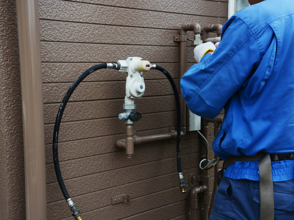 A professional gas technician in a blue uniform inspecting and servicing an outdoor gas pressure regulator and line assembly on a residential home.
