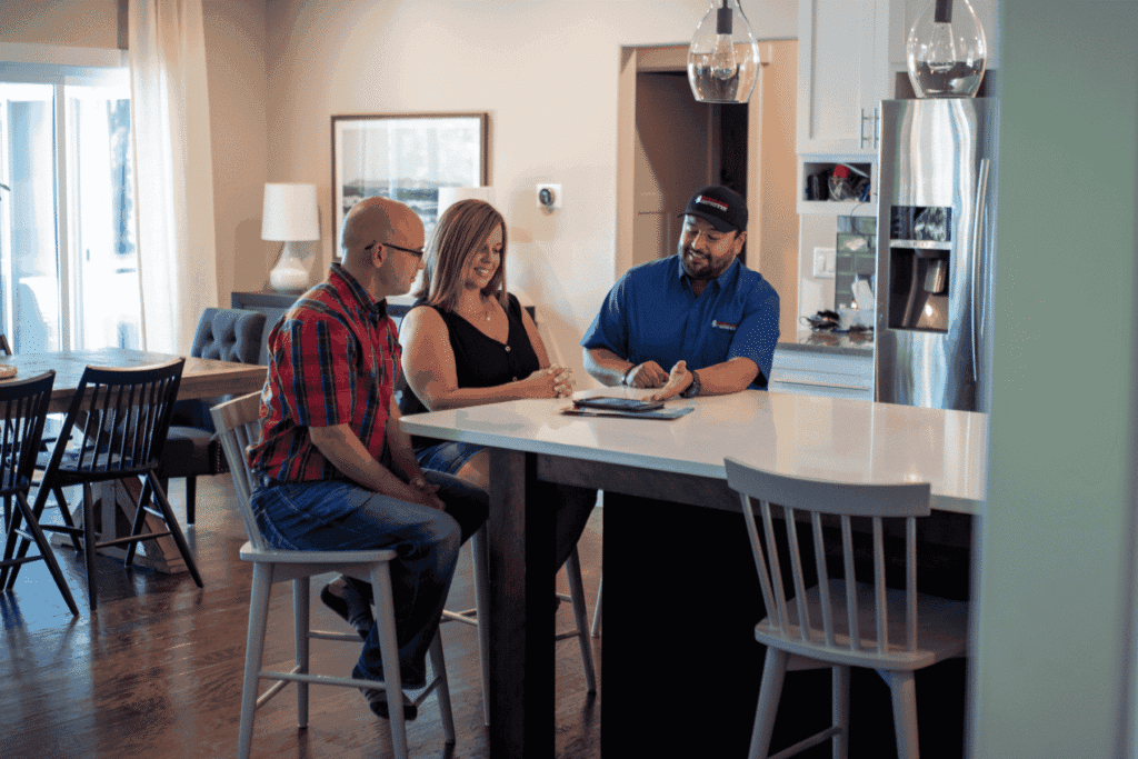 Three adults sit at a kitchen island; a man in uniform shows documents to a seated couple in a modern home setting.