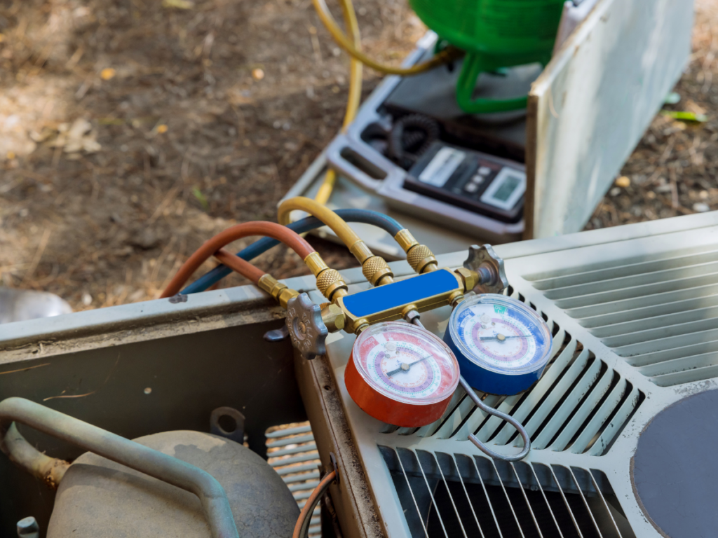 A set of HVAC manifold gauges connected to an outdoor air conditioning unit during maintenance, with a refrigerant tank and digital scale in the background.