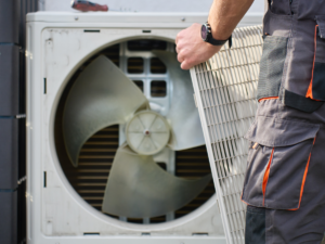 A person in work clothes removes the protective grill from a large outdoor air conditioning unit, revealing the fan and inner components.